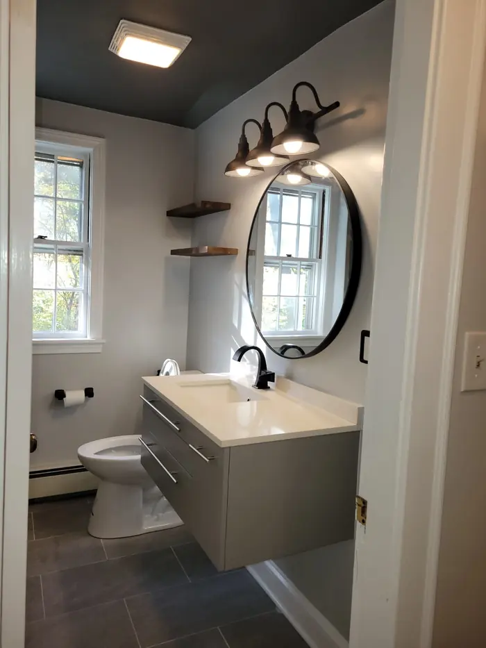 Remodeled bathroom with floating grey vanity, round black mirror, black faucet, walnut floating shelves and dark painted ceiling